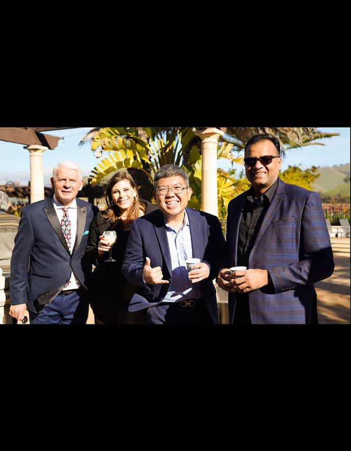 He enjoys a sunny outdoor networking event with three professionals, all holding coffee cups and sharing a cheerful moment.
