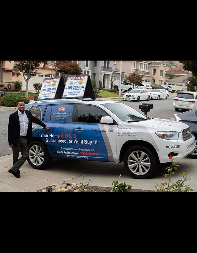 poses beside a branded company vehicle of Your Home Sold Guaranteed Realty, featuring bold advertising promises on a suburban street.