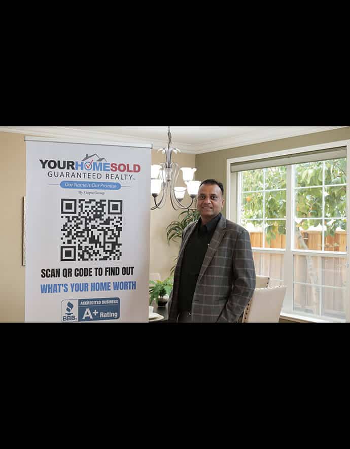 Sharad Gupta stands confidently next to a promotional stand featuring a QR code for Your Home Sold Guaranteed Realty in a well-lit room with garden views.